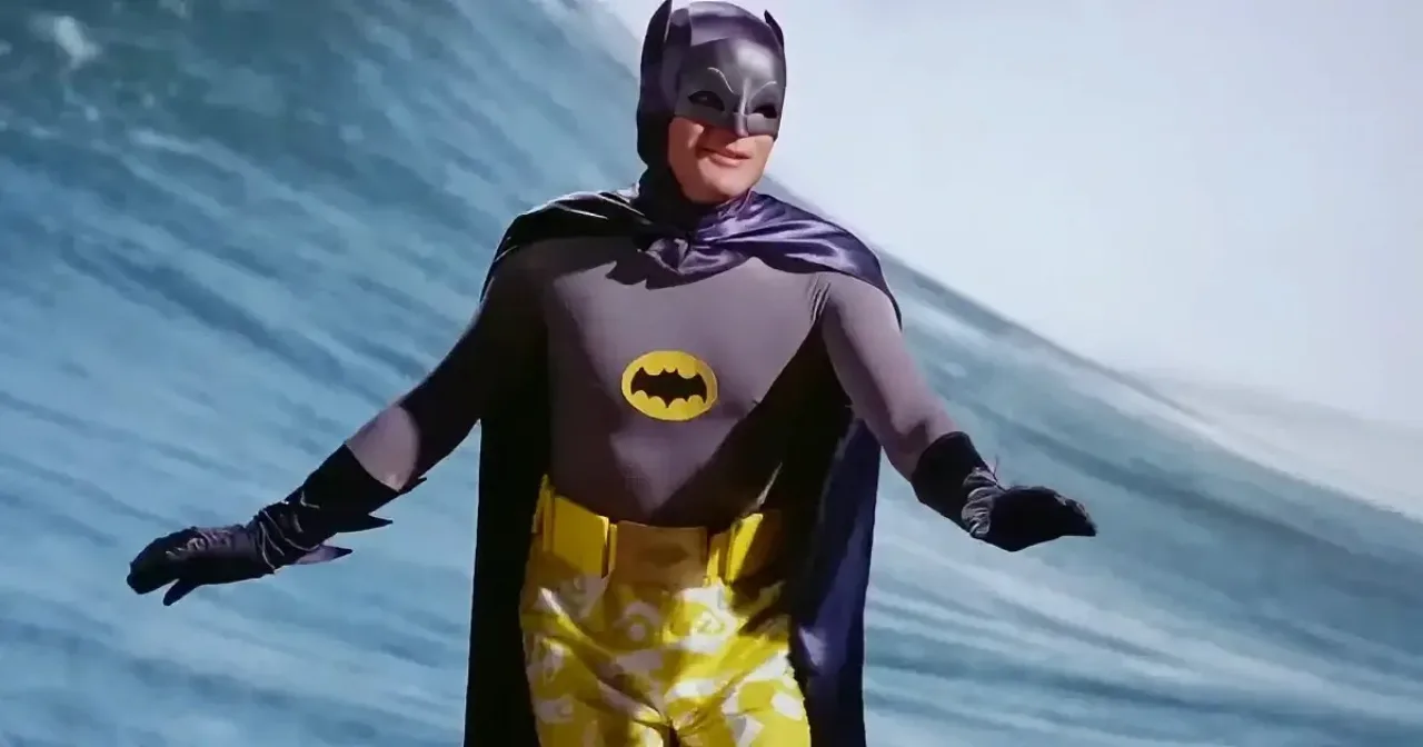 Batman e Coringa em trajes de 1966 surfando na praia, com bat‑rockets ao fundo e cores pop‑art vibrantes