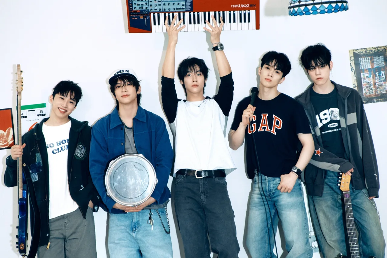 Membros de N.Flying posam em traje escuro ao lado da capa do mini‑álbum “Still” com o logo da banda