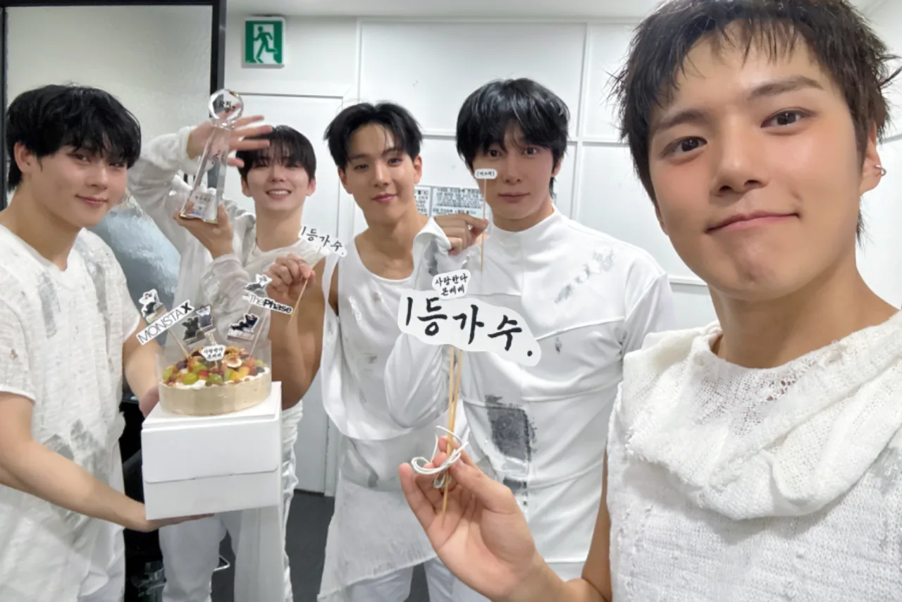 MONSTA X no palco de Music Bank, iluminado, cantando “MAGIC” com microfones e o troféu ao fundo