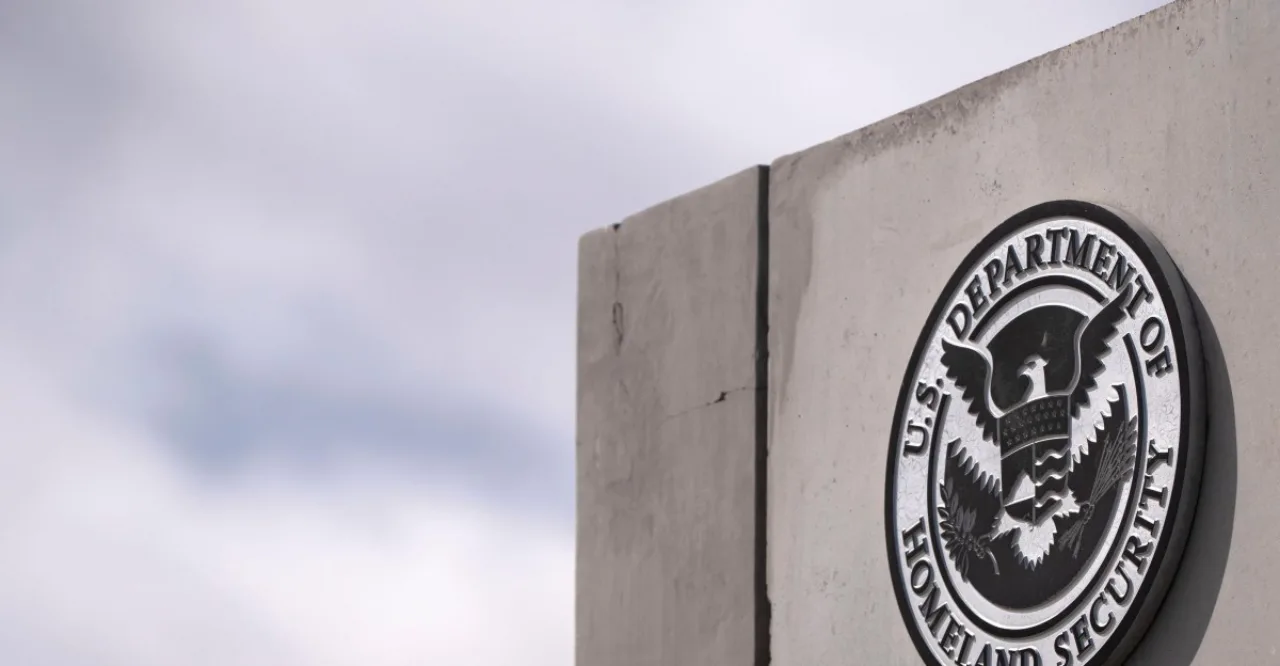 Silhueta de hacker diante de monitor exibindo logotipo do DHS e planilhas de dados expostos