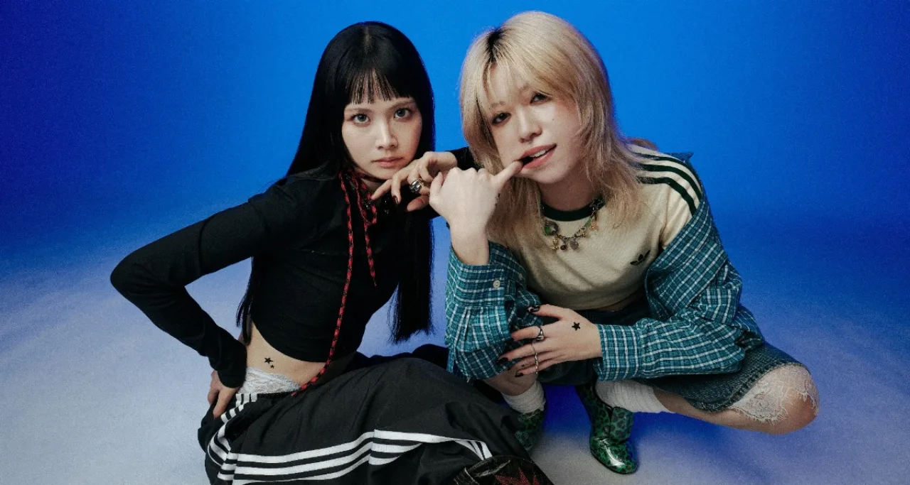 Emma e Menu, do duo EMNW, posam com microfones ao lado da capa do single “LOVING TO GET US BY”