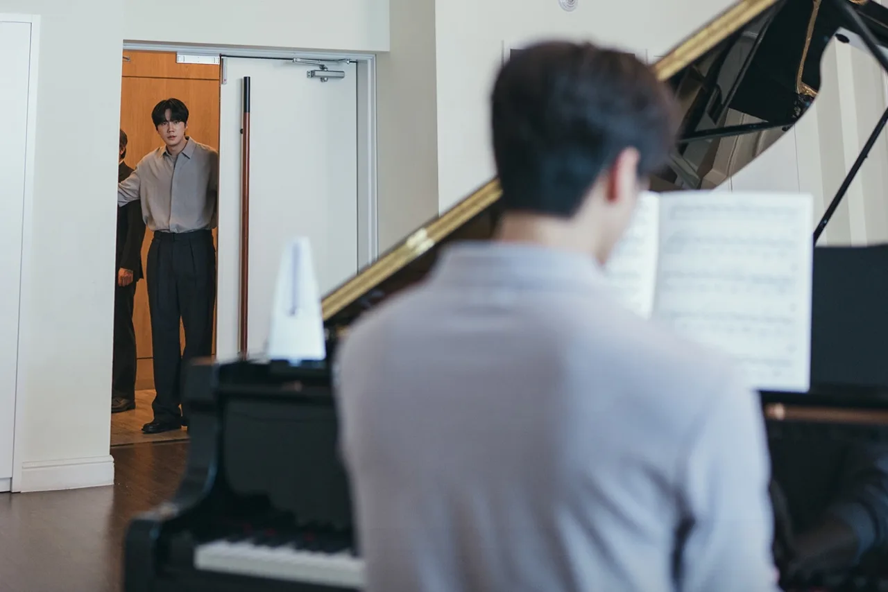 Song Kang, de terno, ao piano, encarando Lee Jun Young, que observa ao fundo, no palco de concerto